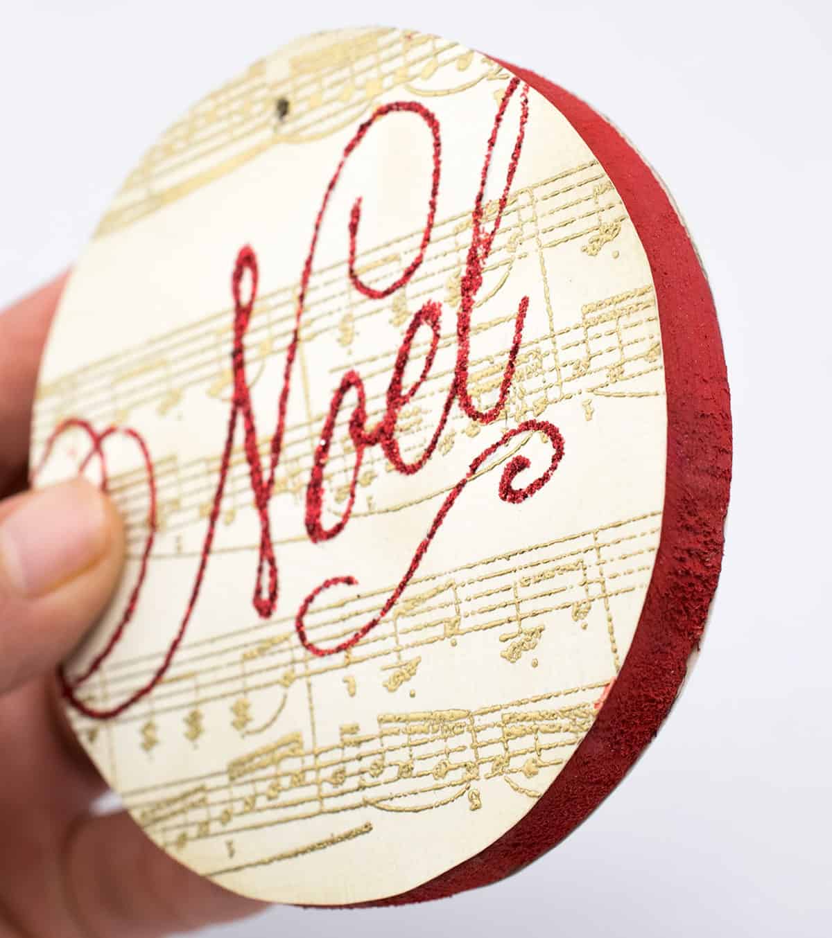 DIY stamped ornament on wood disk with gold sheet music and red glitter noel image with red painted sides. 