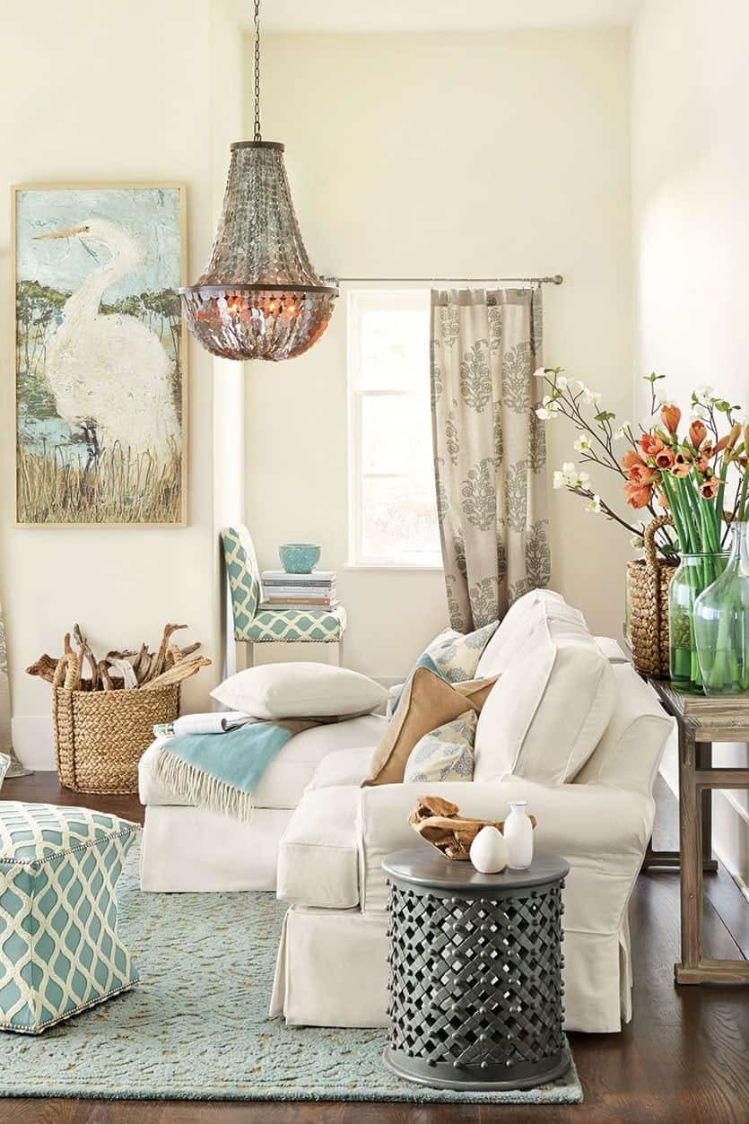 Beige sofa on soft green rug in living room with jewel chandelier, green chair and footstool, metal side table, flowers, and pelican painting. 