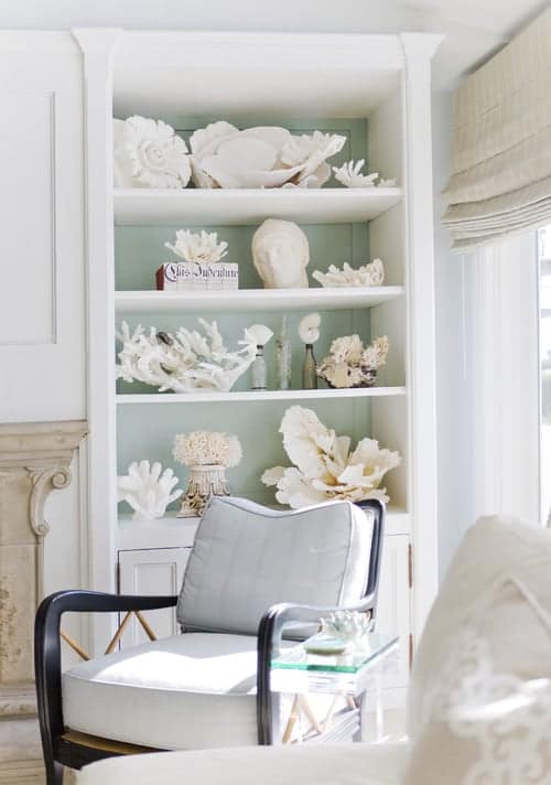 Large white Shelf with collection of white coral with soft green background with gray chair in front. 