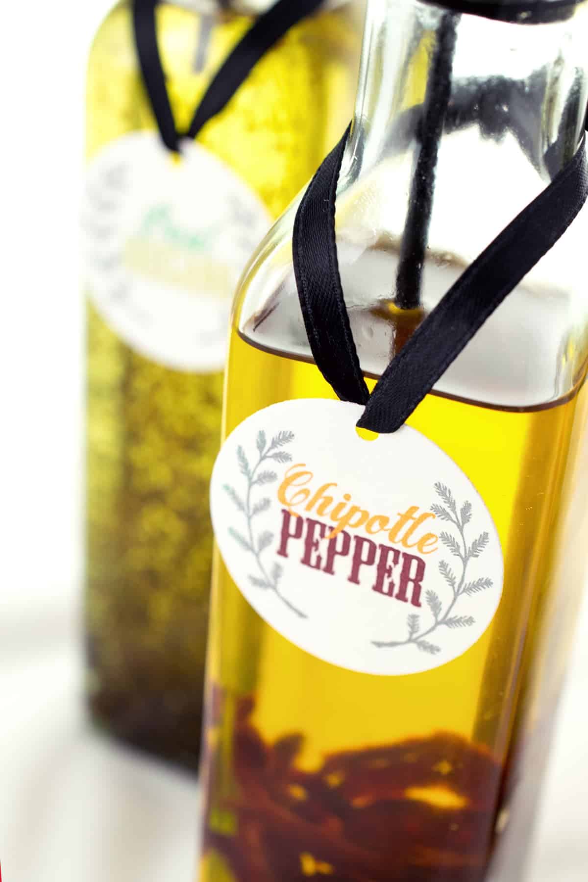 Closeup of printed chipotle pepper label tide to bottle of infused oil with black ribbon with basil oregano oil in background.