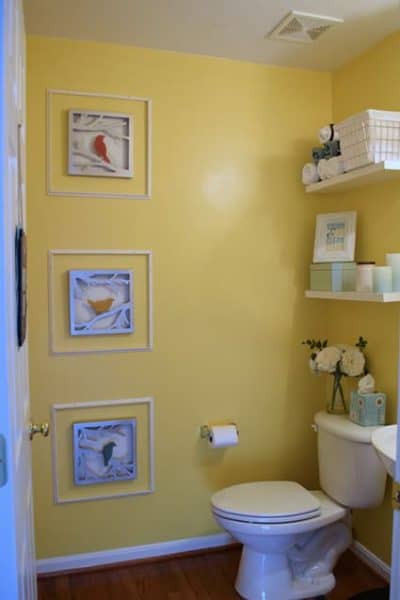 Small yellow bathroom with white commode, white floating shelves, and three framed 3-D bird art. 