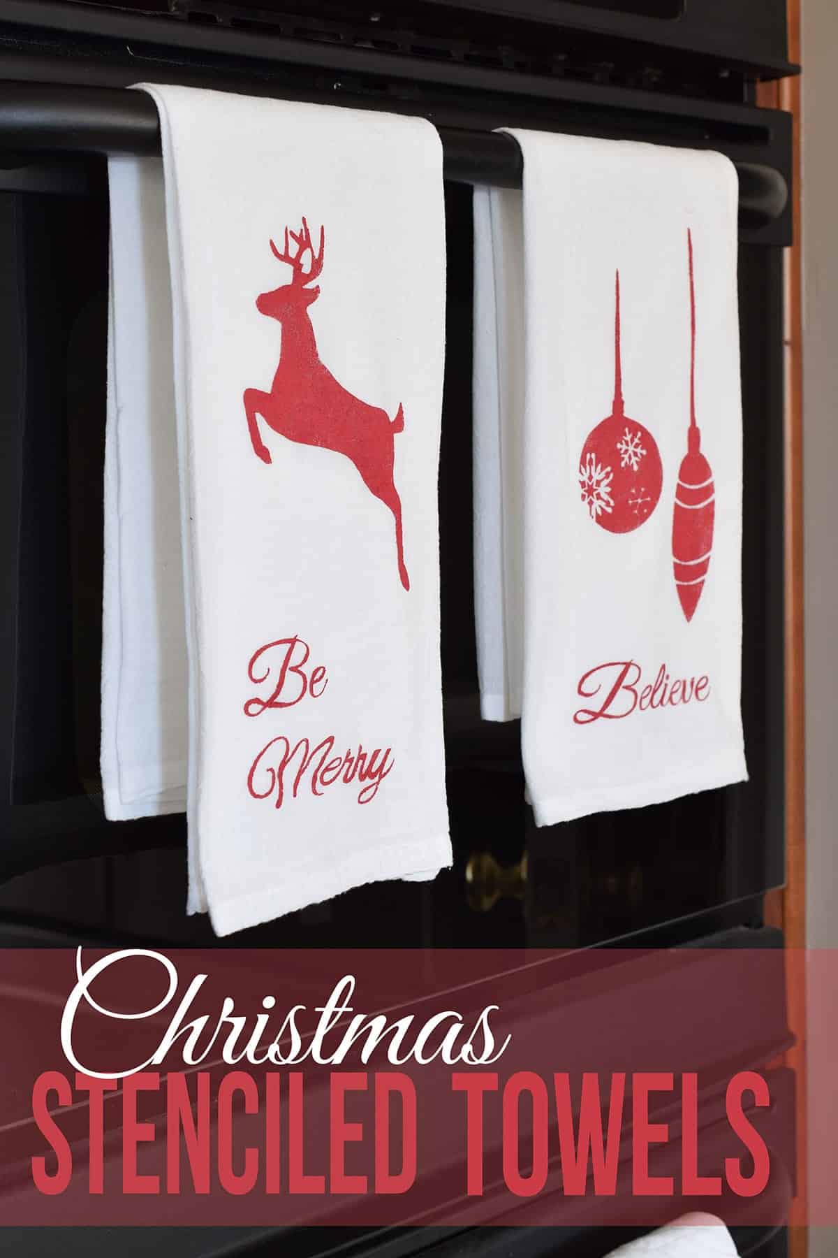 Two white Christmas themed tea towels with red deer and ornament stencils hanging on black oven door with post title. 