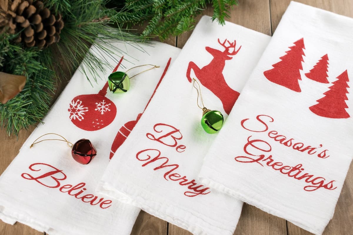 Three white tea towels with red Christmas themed stencils spread on wooden surface with garland, pine cones, and ornaments. 