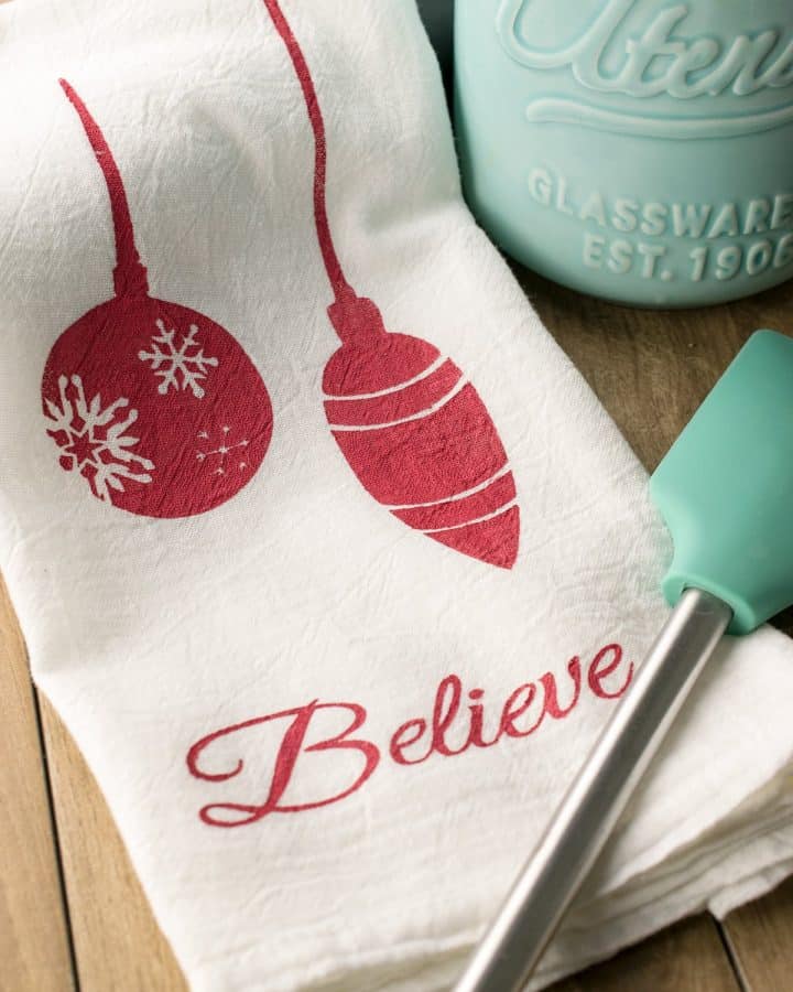 Christmas Stenciled Tea Towels laid on a table jumping reindeer, ornaments, Christmas Tree silhouettes, and text.