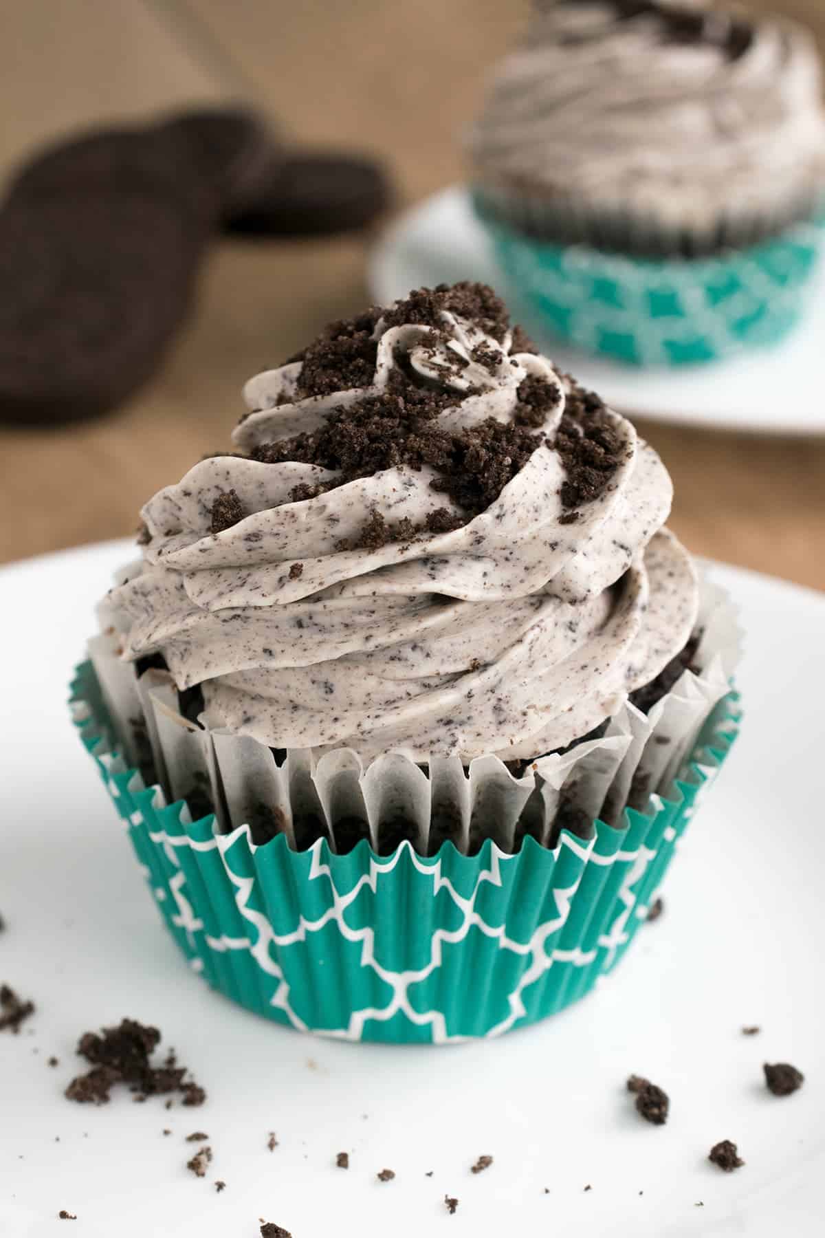 Oreo cookie frosting swirled on top of chocolate cupcake in turquoise liner on white plate, dusted with Oreo crumbles.