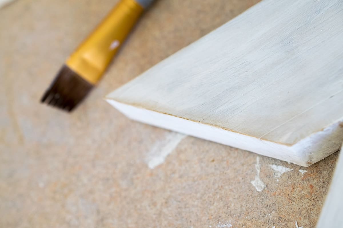 Corner of dry brush painted stained wood with white chalk paint next to paint brush. 