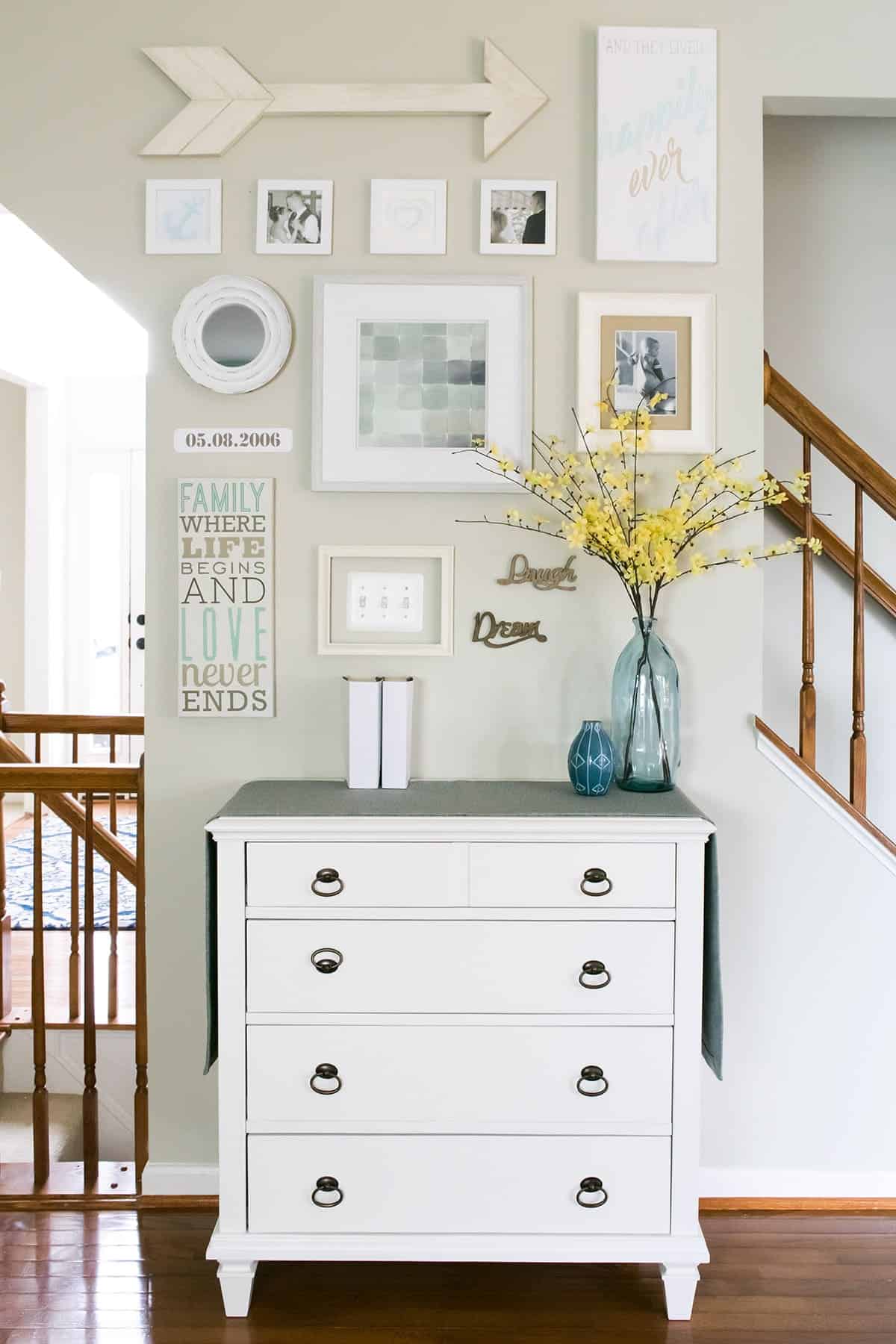 Wall art gallery with DIY wooden arrow, DIY art and frames above white chest of drawers with glass vase filled with bright yellow flowers. 