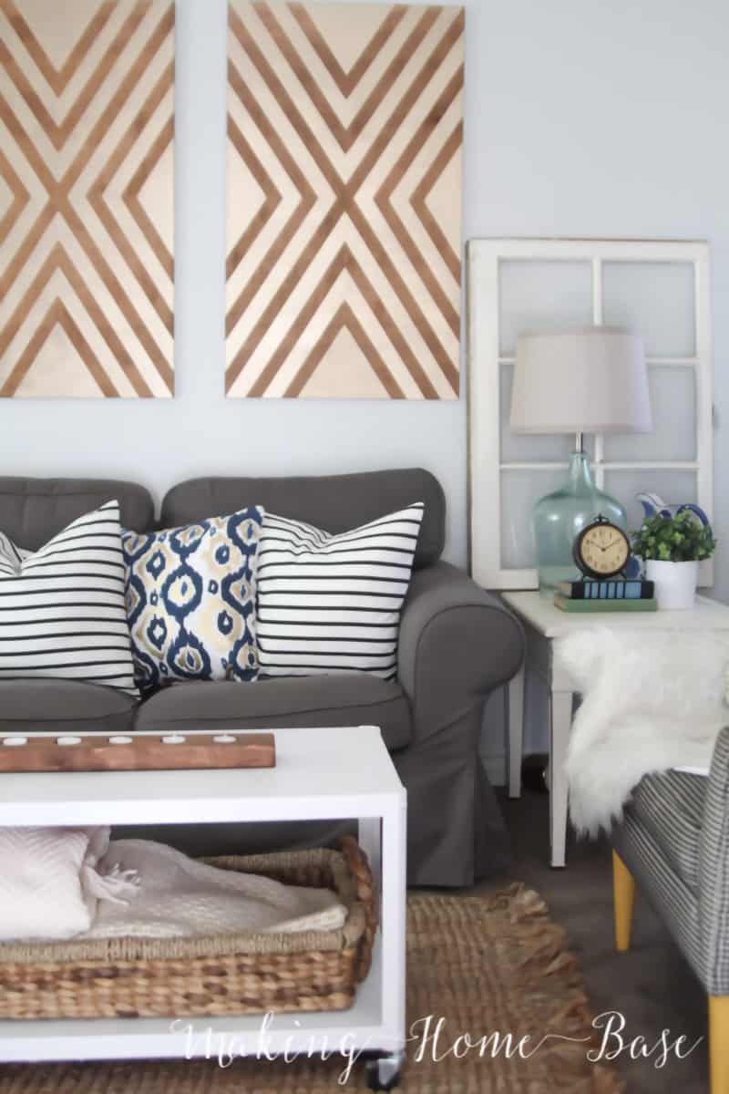 Geometric wood panel wall art in room with gray couch, white and blue accent pillows, and white table with wicker basket and wood centerpiece. 
