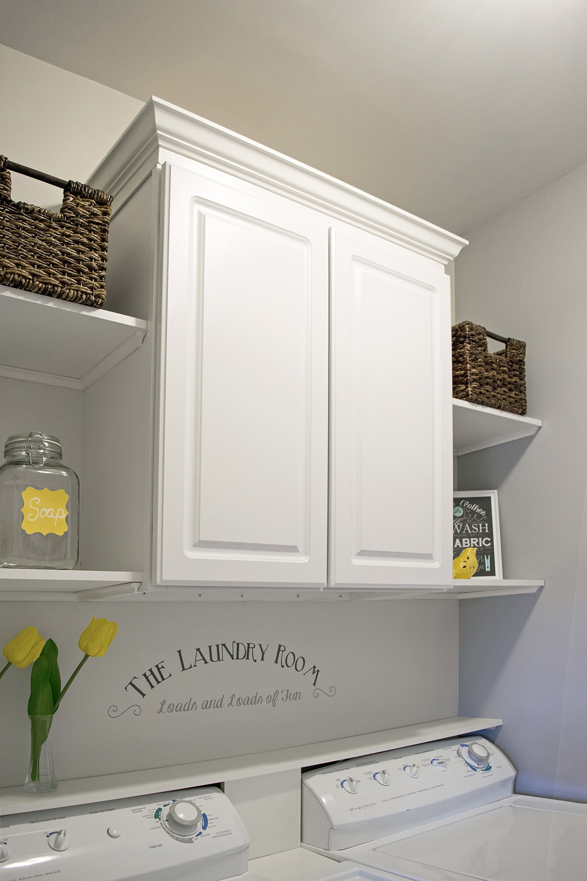 Installed cabinet and shelves in laundry room with decorative baskets and yellow flowers above washer and dryer. 