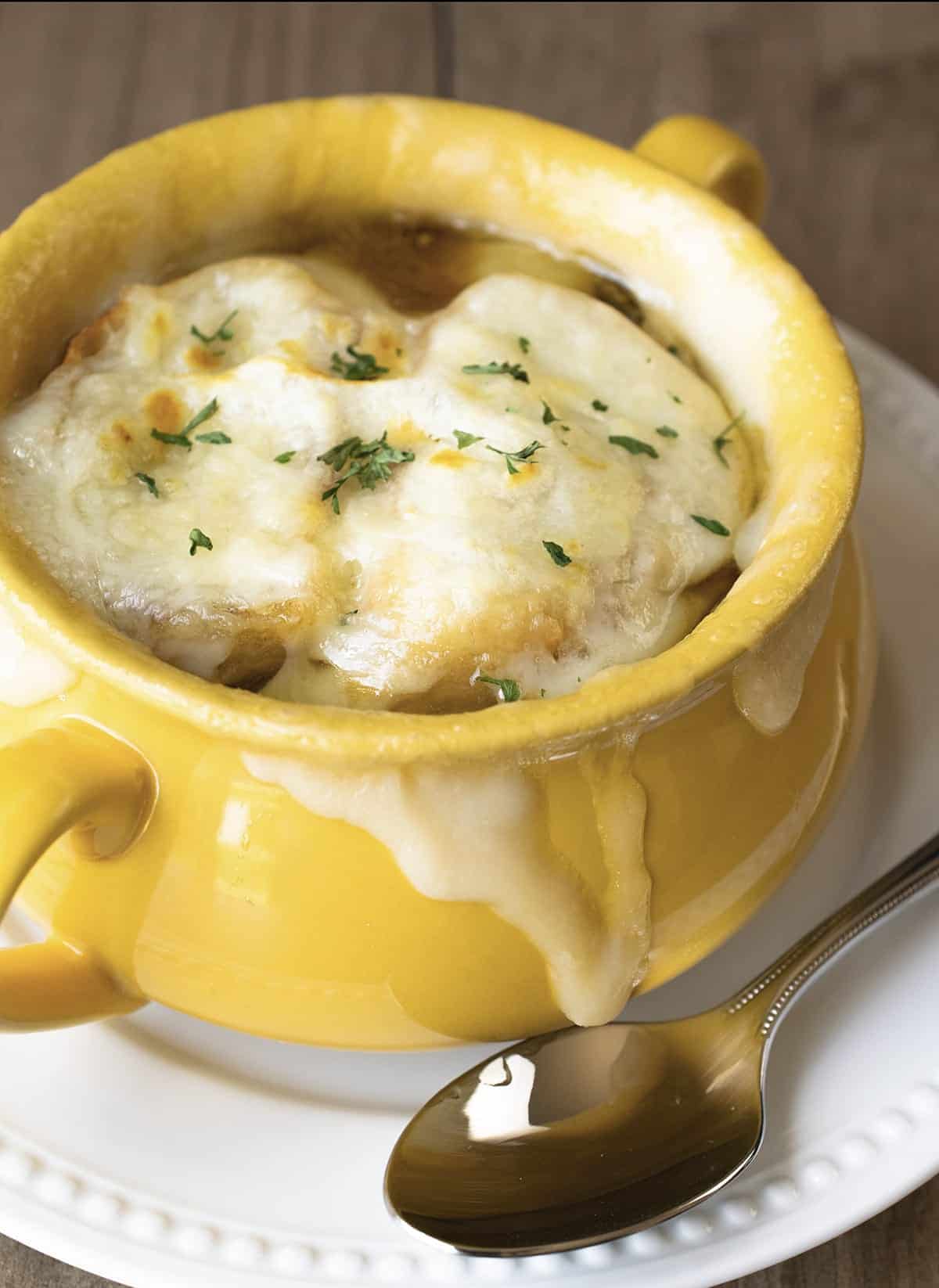 White plate and spoon with yellow bowl of french onion soup topped with melted cheese and parsley. 