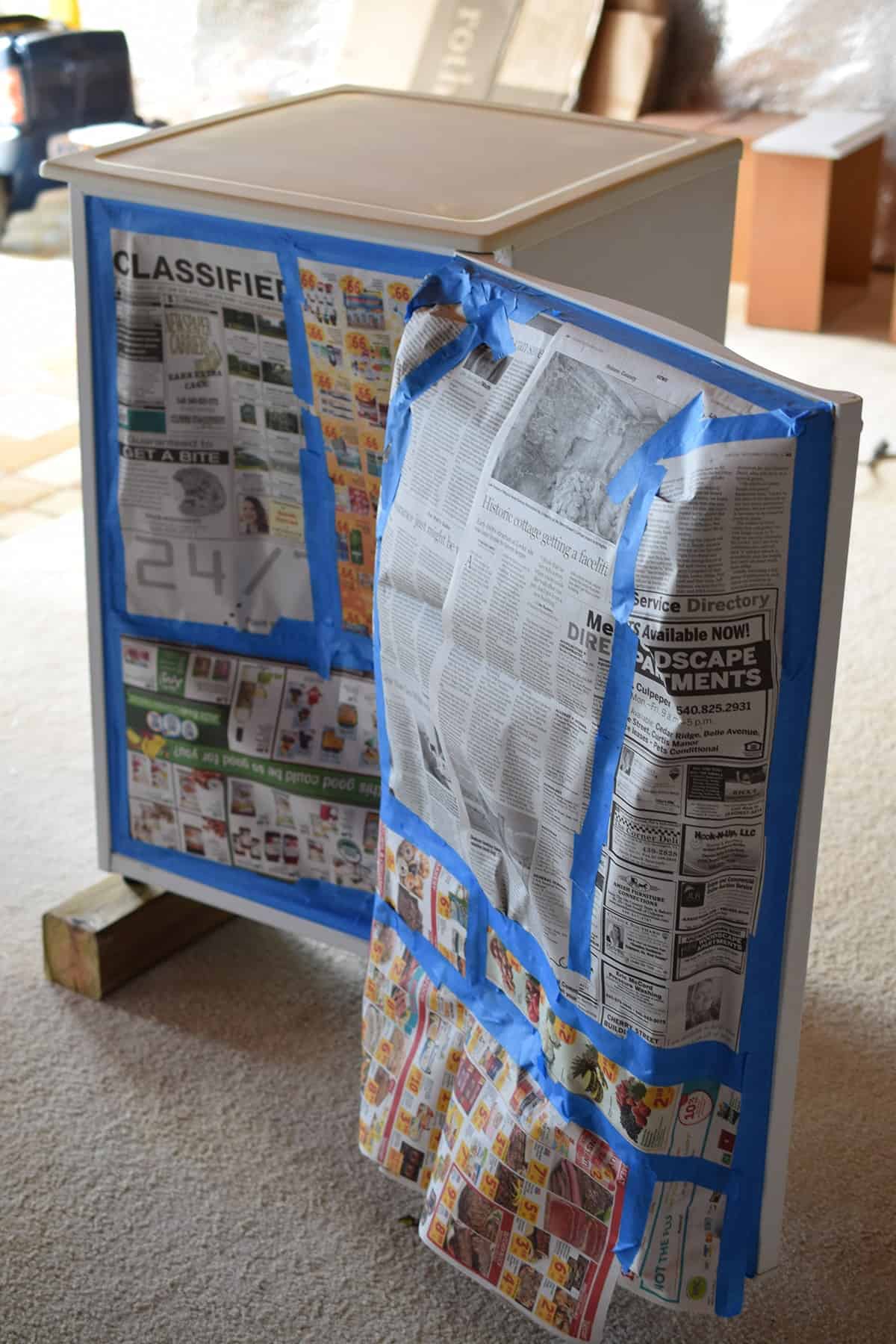 White mini fridge on wooden blocks, taped with newspaper in preparation for painting. 