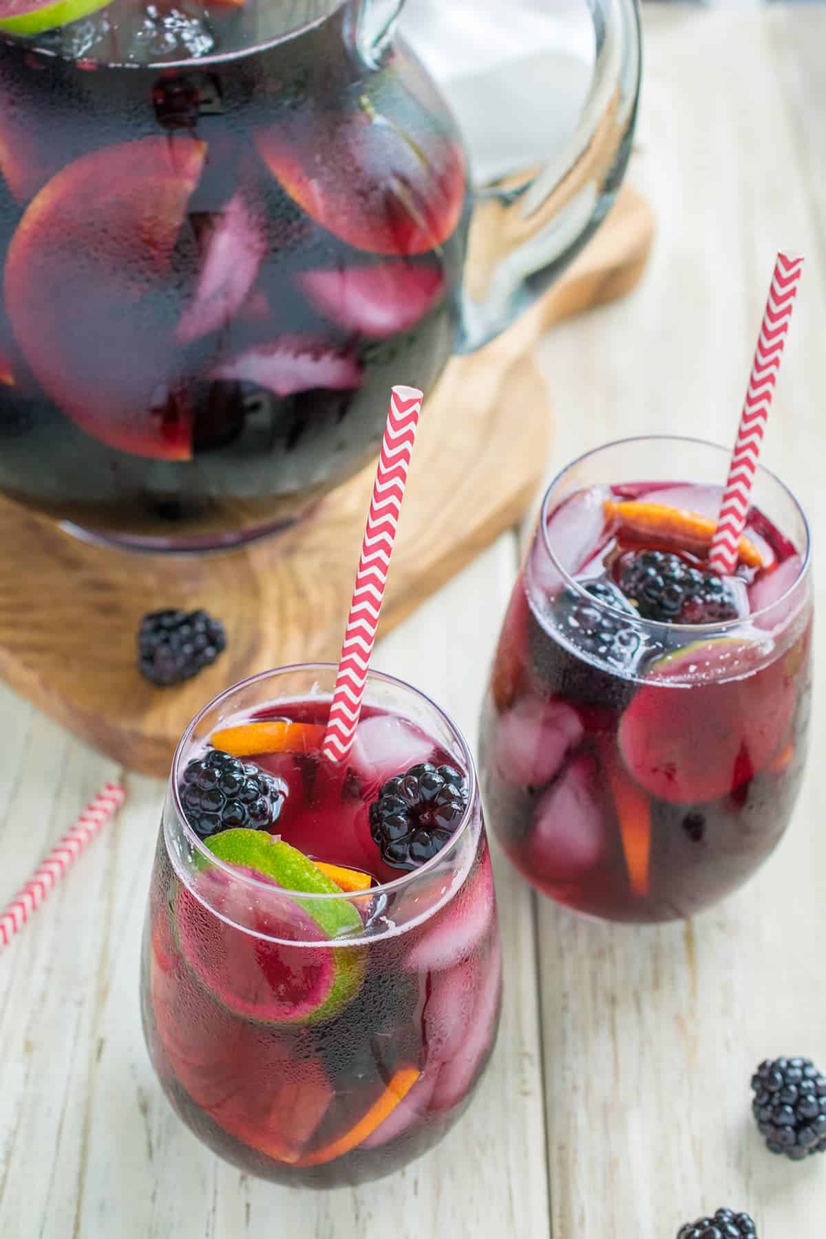 Glass pitcher of blackberry sangria on wooden paddle next to two wine glasses will with sangria and fresh sliced fruit.