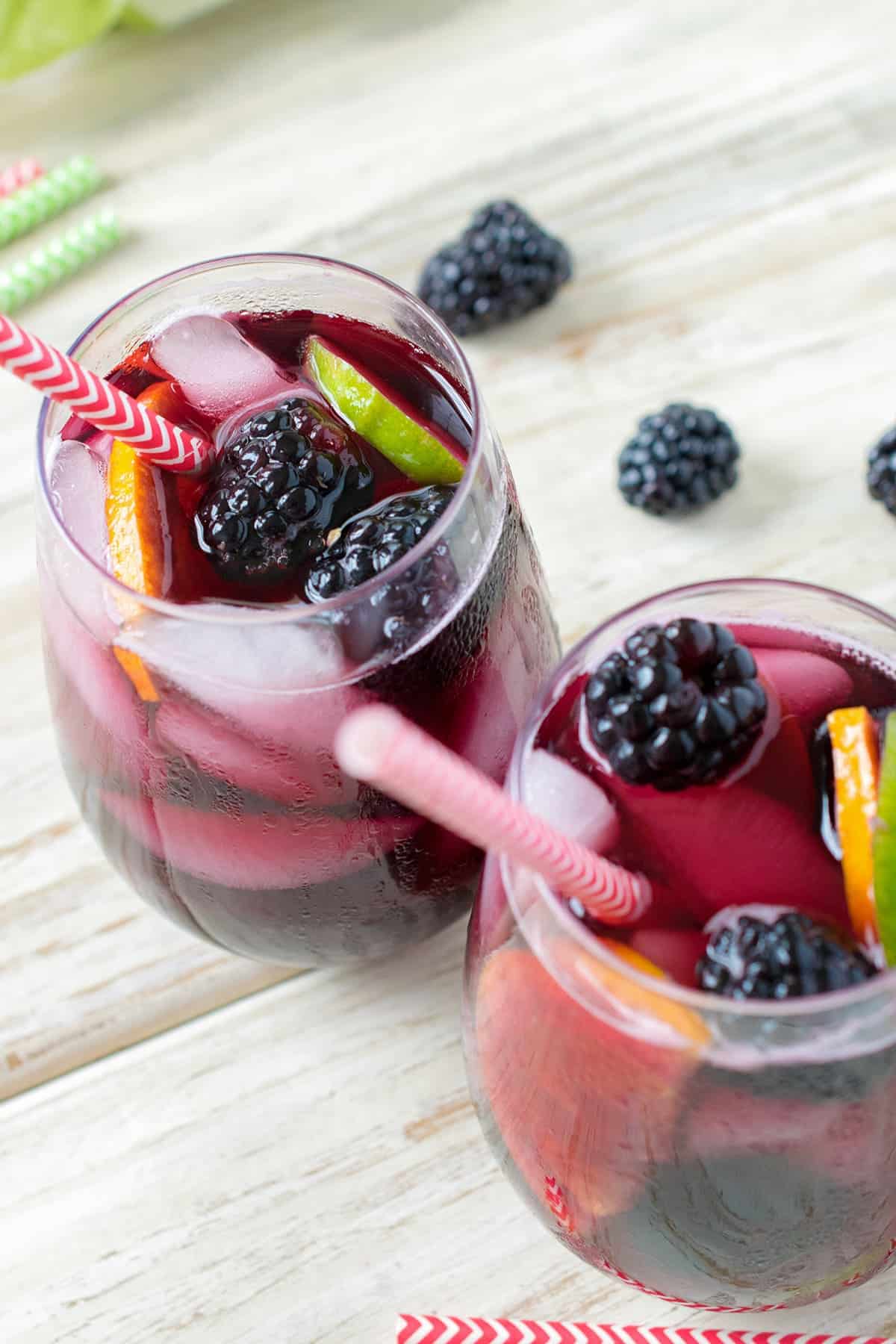 Stemless wine glasses filled with sweet blackberry sangria, over ice, with fresh fruit and red and white straws. 