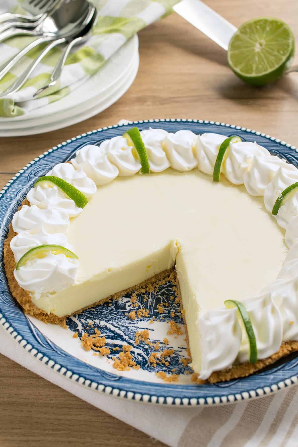 Whole lime pie in blue dish with one slice removed. Stacked white dishes with folded napkins and silverware 