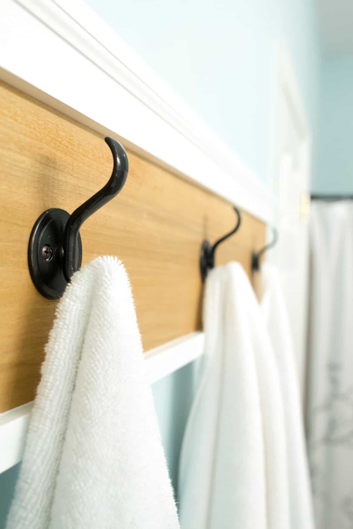 Natural wood towel rack with white molding and black metal hooks mounted on bathroom wall. 