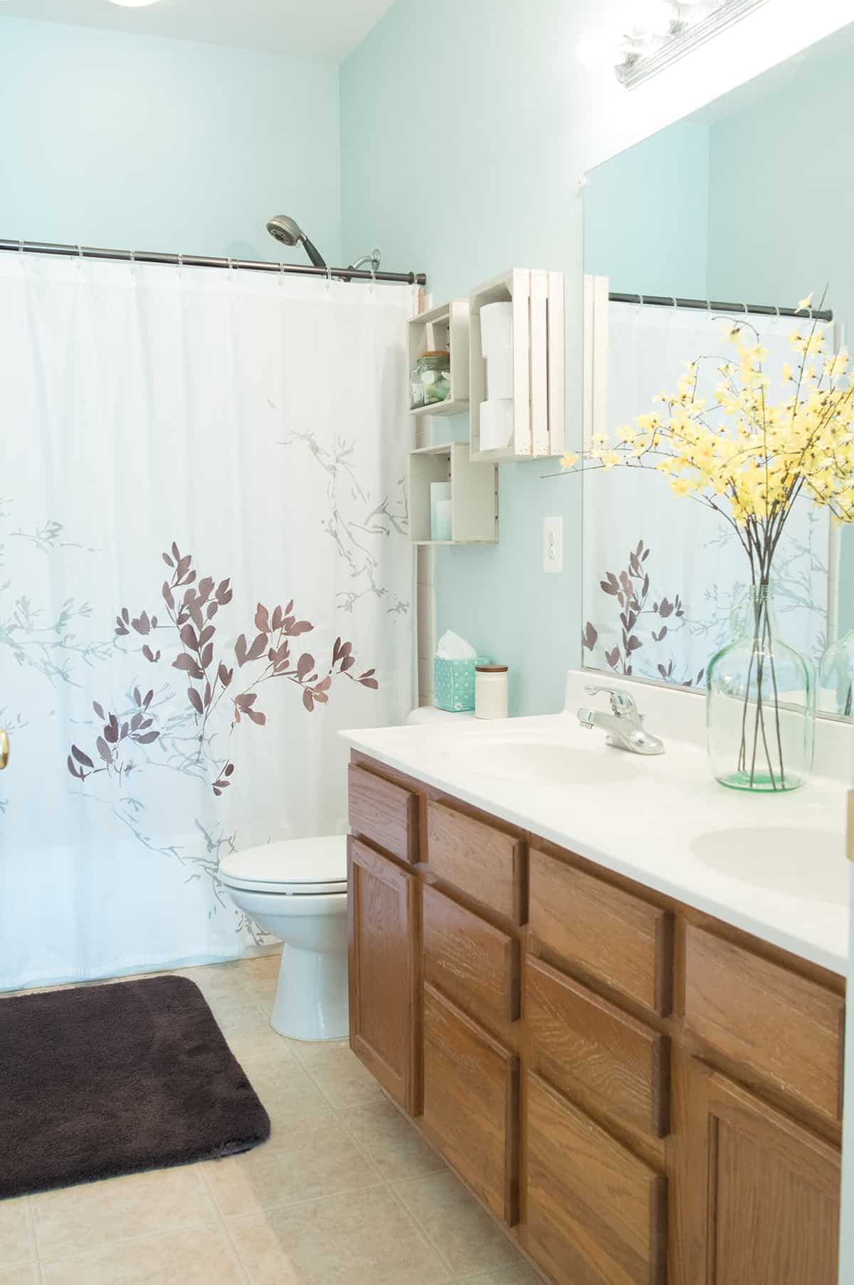 Spa inspired bathroom with floral accents, soft bluish-green wall color, and added wooden crate shelves above commode. 