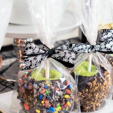 Chocolate covered apples rolled in crushed candy, wrapped in clear bags with black ribbon on white platter.