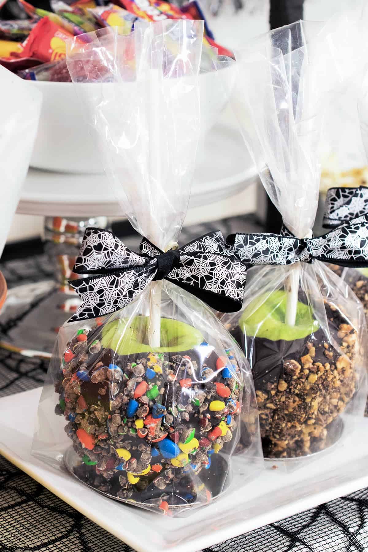 Chocolate covered apples rolled in crushed candy, wrapped in clear bags with black ribbon on white platter. 