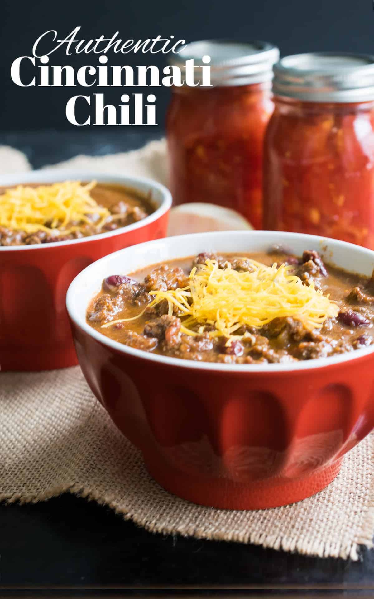 Hearty Cincinnati style chili in red bowls with mounds of shredded cheese next to mason jars of tomatoes. 