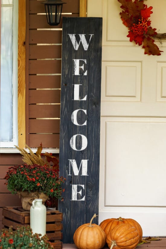 Black letter porch sign with welcome printed on it and surrounded by pumpkins.
