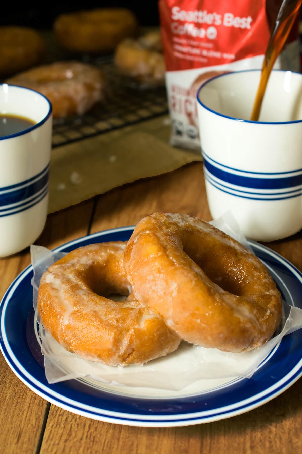 Two glazed old fashioned sour cream doughnuts stacked on white and blue plate next to cups of black coffee. 