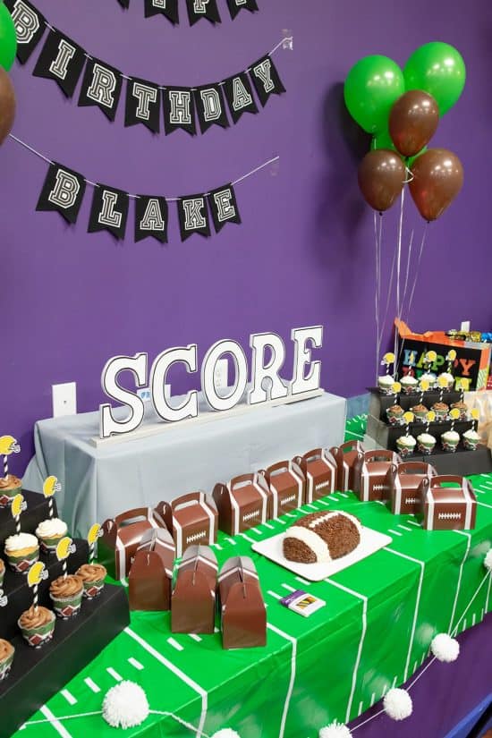 Front shot of sports-themed party table laid with green field patterned tablecloth, white pom garland, football cake & tiered cupcakes and football shaped party favor boxes with white SCORE letters in the background.