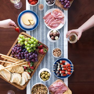 Overhead of a cheese and wine party setup with foods scattered on the table and hands moving to pick up items.