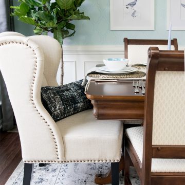 Traditional dining room reveal features plush linen wingback chair with brass tack detail and dark legs accented by modern black and white throw pillow. Traditional dining table and chairs sits on black and white floral pattern oriental rug covering dark finished floors. Light wainscotting leads up to blue-green patterned walls with framed white matted bird prints.
