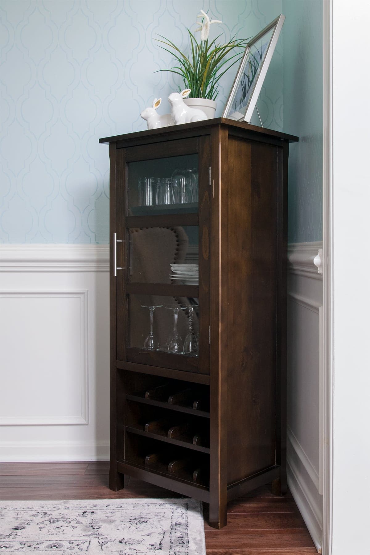 Dark wood wine cabinet in corner of light blue dining room with potted plant, framed photo, and white decorative rabbits. 