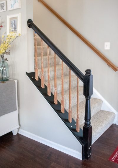 Bannister painted black with unfinished spindles and unfinished handrail parallel with light carpeted steps coming off dark hardwood floors and light gray walls.