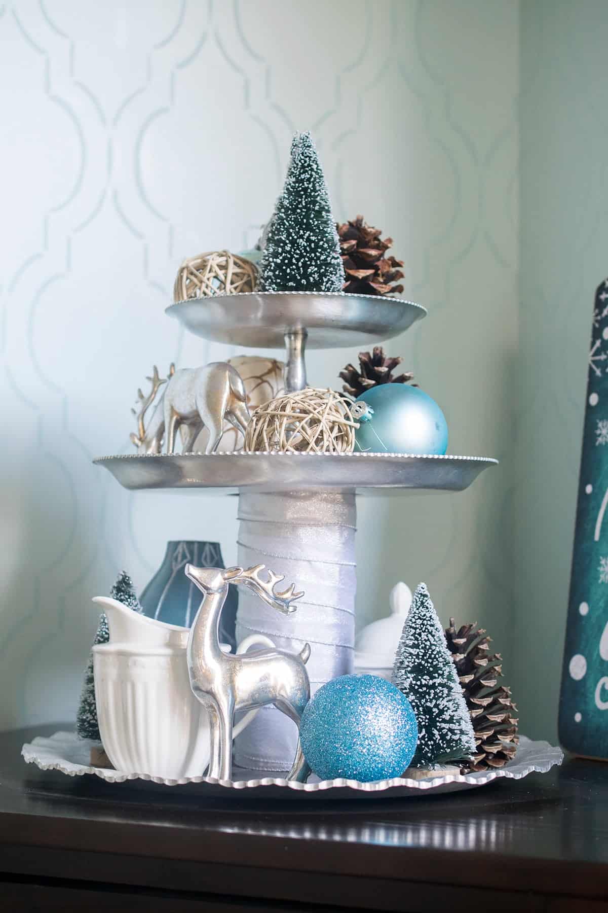 Tiered tray decorated for Christmas with silver deer, pinecones, blue ornaments, and bottle brush trees.