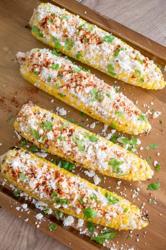 Four elotes or ears of corn dressed in mexican cheese and spices laying on a table top.