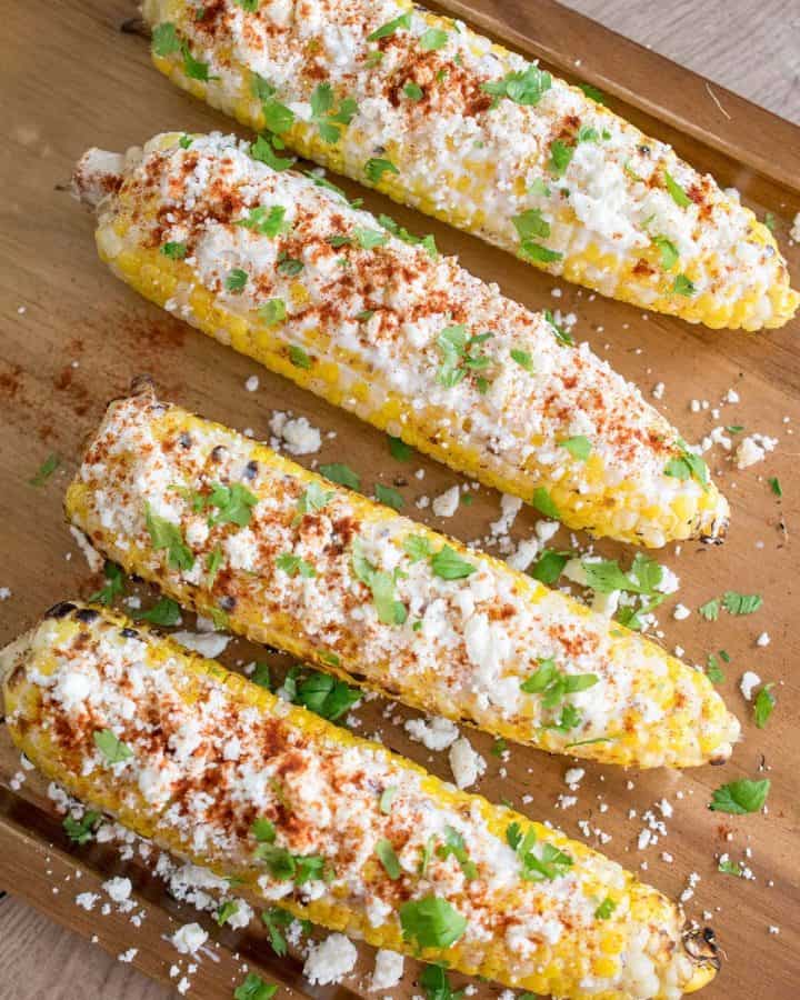 Four elotes or ears of corn dressed in mexican cheese and spices laying on a table top.