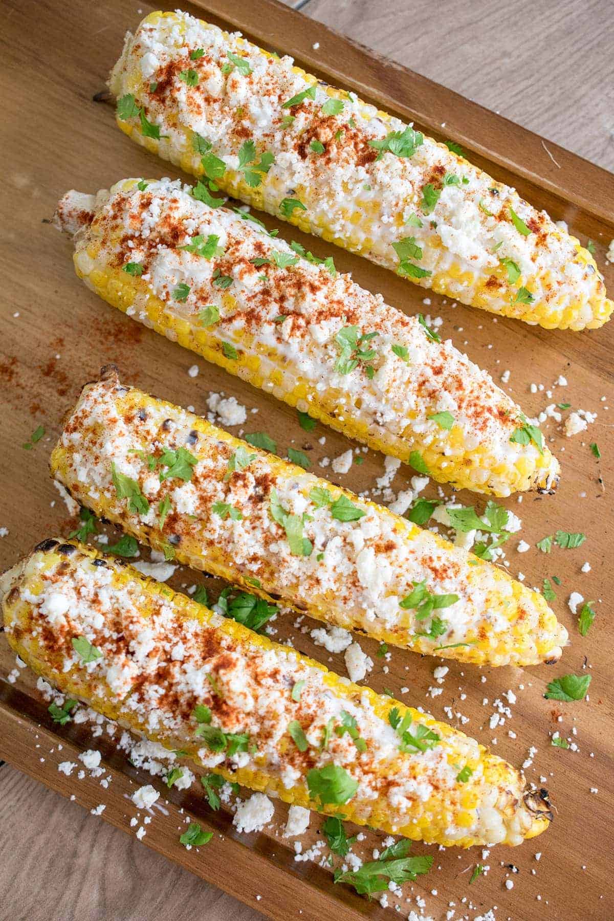 Four elotes or ears of corn dressed in mexican cheese and spices laying on a table top.