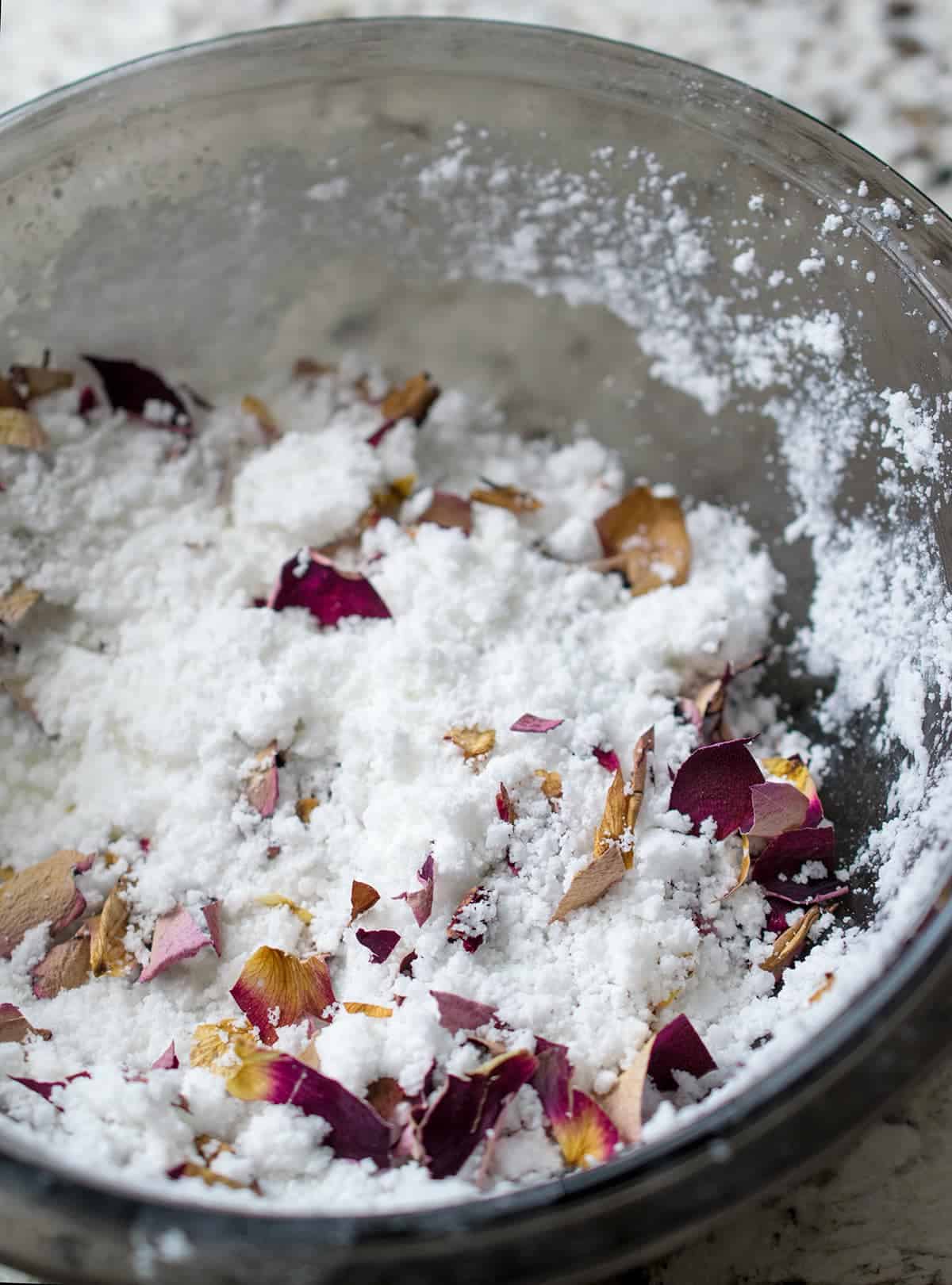 White bath bomb base mixture with added dried rose petal pieces in glass bowl