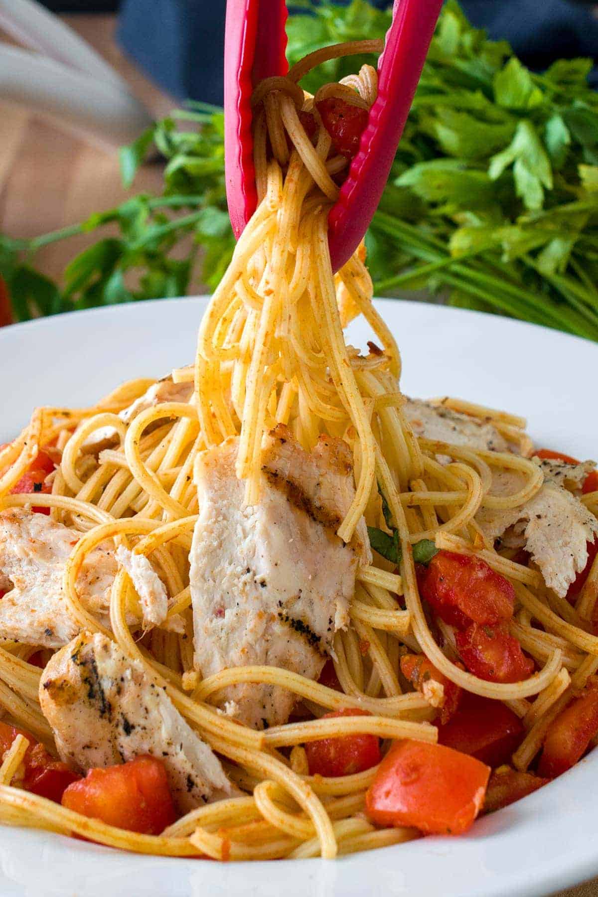 Tongs picking up bruschetta chicken pasta with sliced chicken, tomatoes, basil, and parmesan falling to the side.