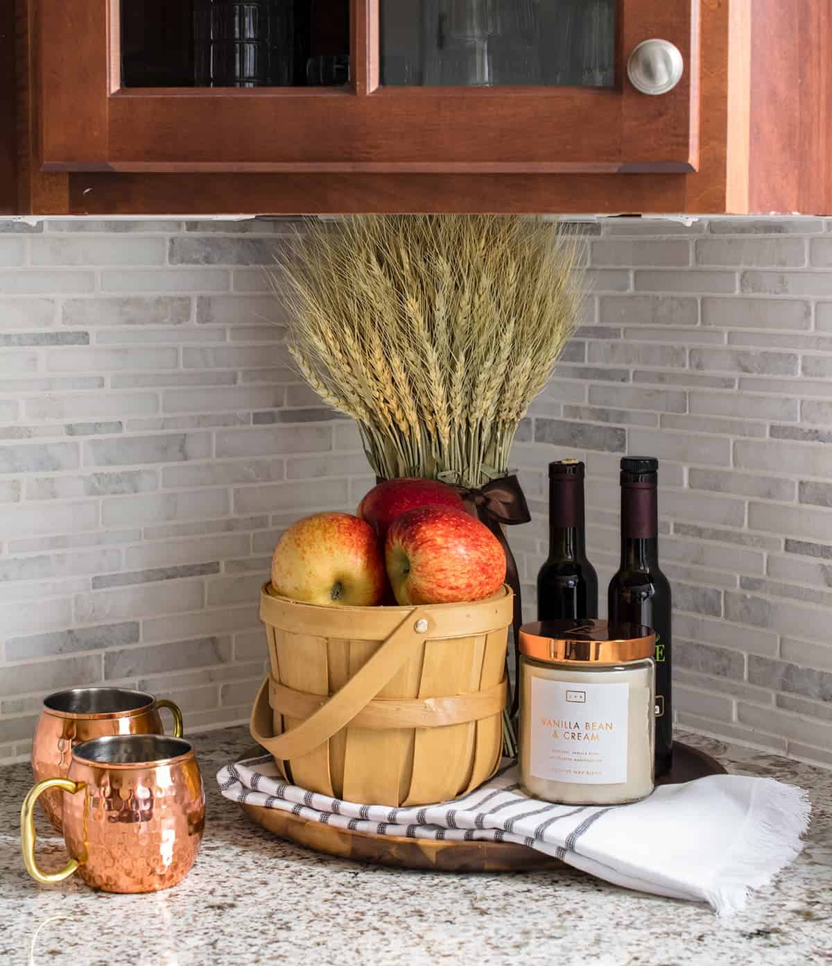 Fall accented kitchen tray with wooden pale filled with apples, artificial grains, candle, and copper mugs. 