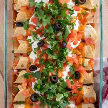 Top down view of baked chicken enchiladas with sour cream, pico de gallo, olives, and cilantro as garnish in glass baking dish.