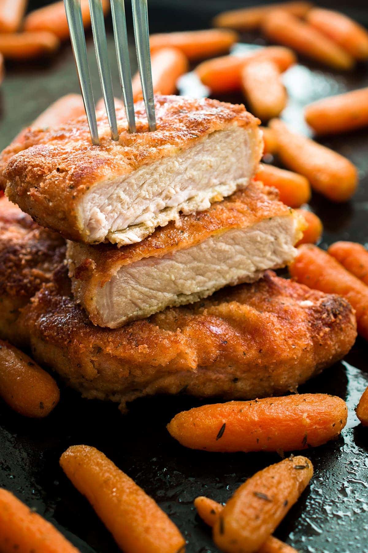 Breaded baked pork chops with baby carrots. Top pork chop sliced in half with fork piercing through it.