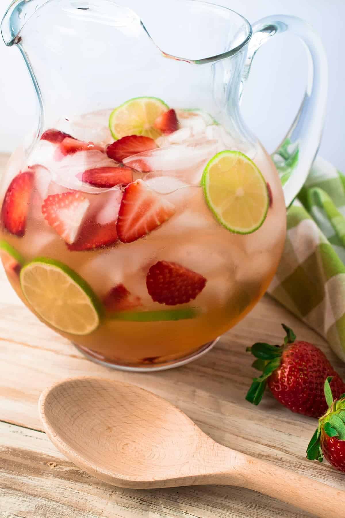 Glass pitcher of strawberry sangria with wooden spoon and whole strawberries on table.