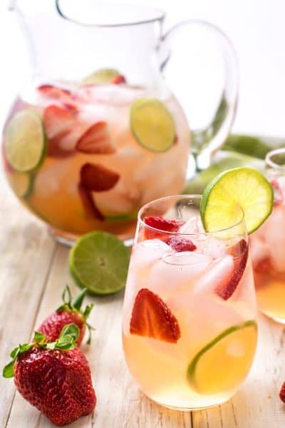 Sangria with strawberries and limes in a glass on a table with a pitcher in the background.