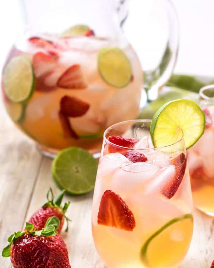 Sangria with strawberries and limes in a glass on a table with a pitcher in the background.