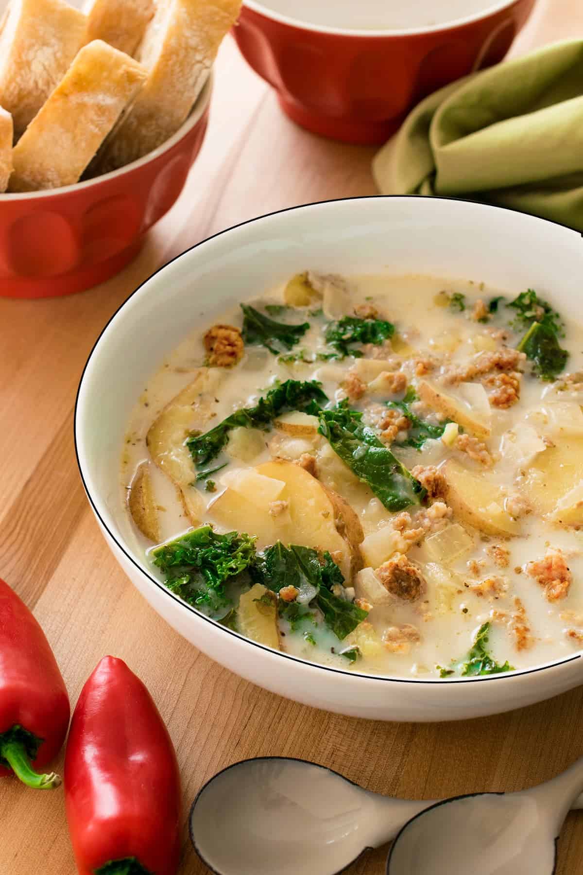 Sausage Kale Soup in large white soup bowl. Red peppers and spoons on the forefront and red bowl of crusty bread to the rear.