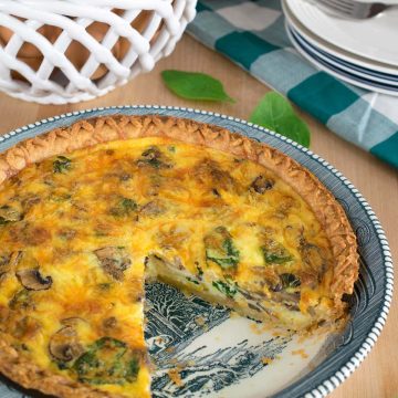 Spinach and mushroom quiche with crust in pie plate with one missing piece. Basket of eggs and stack of plates in background.