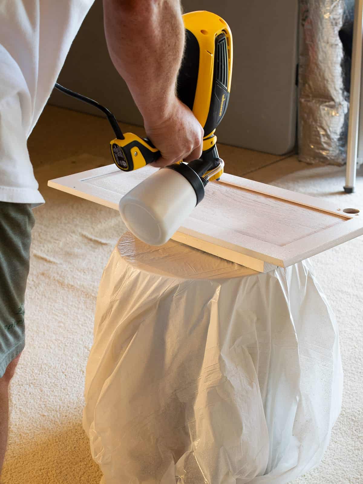 Man using a paint sprayer to paint kitchen cabinet door on raised plastic covered table.