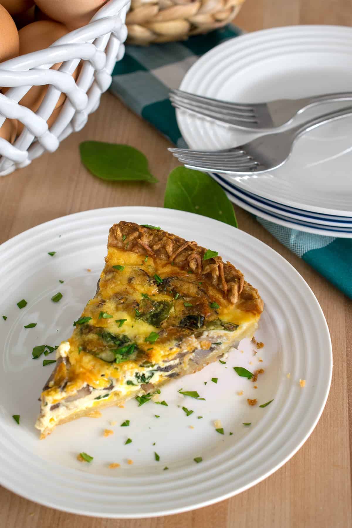 Spinach and mushroom quiche slice on white plate with stack of plates and two forks behind the main feature.