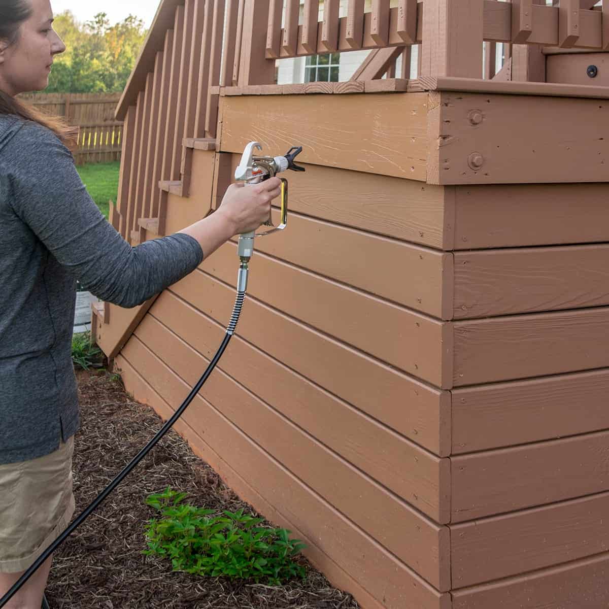 Woman using an airless paint sprayer to paint the wall of a raised deck.