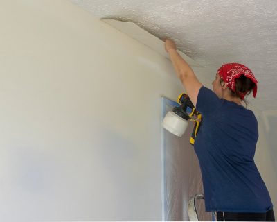 Person painting a wall indoors with a paint Sprayer and holding carboard to edge the paint.