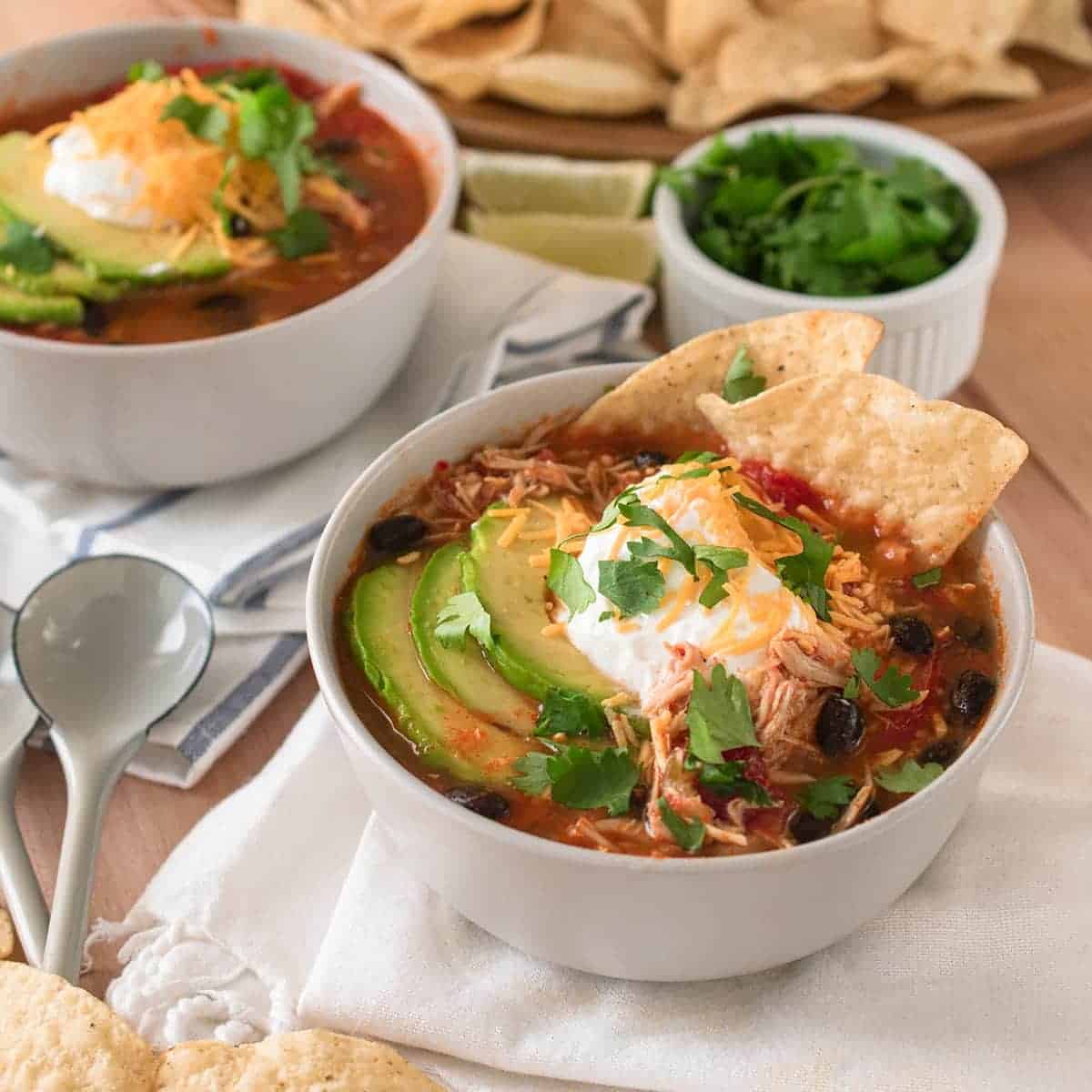 Tortilla soup with toppings of avocado and sour cream. Spoons, a second bowl, a bowl of cilantro and tortilla chips in background.