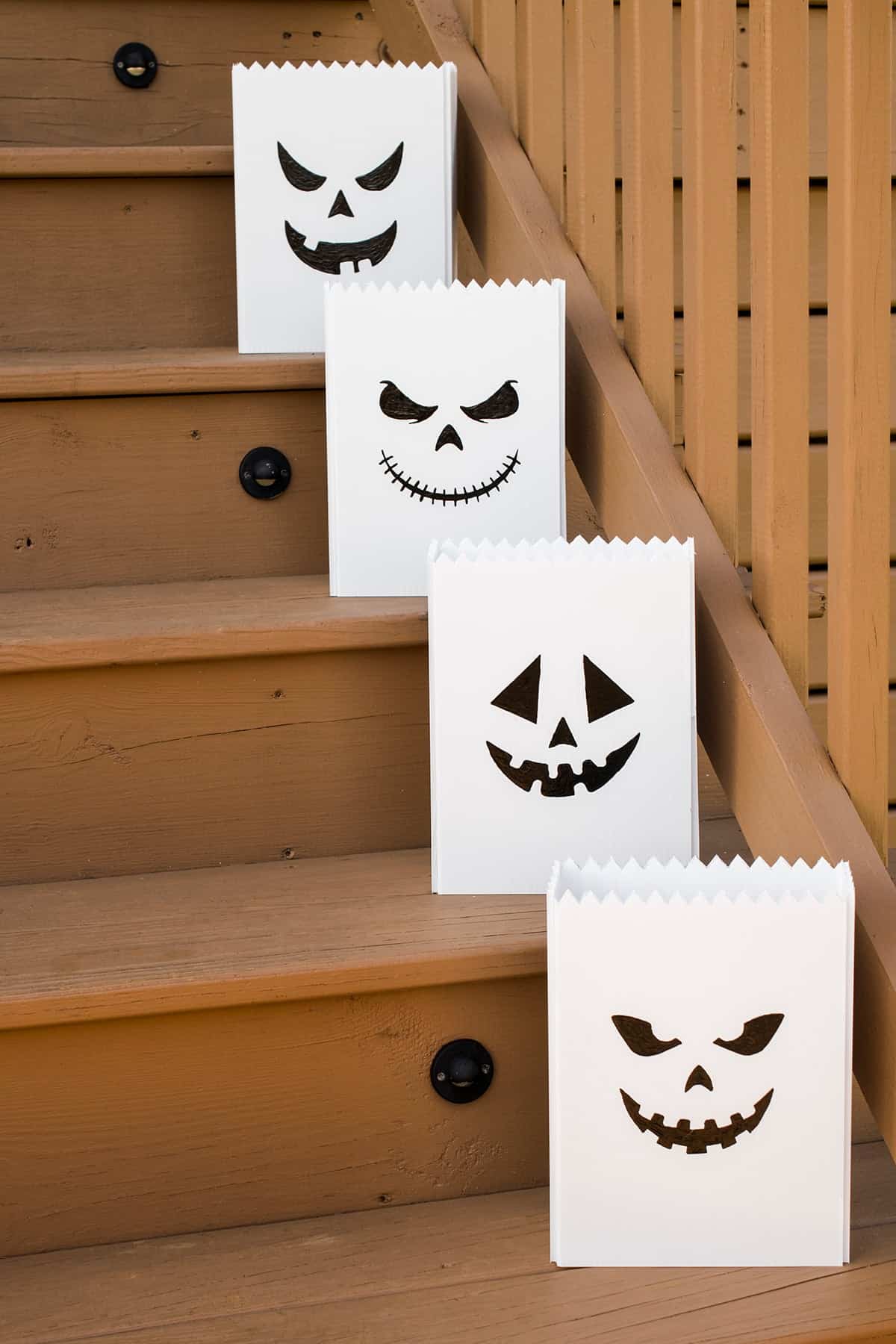 White outdoor pumpkin luminaries with black jack-o-lantern faces on wooden deck steps in daylight.